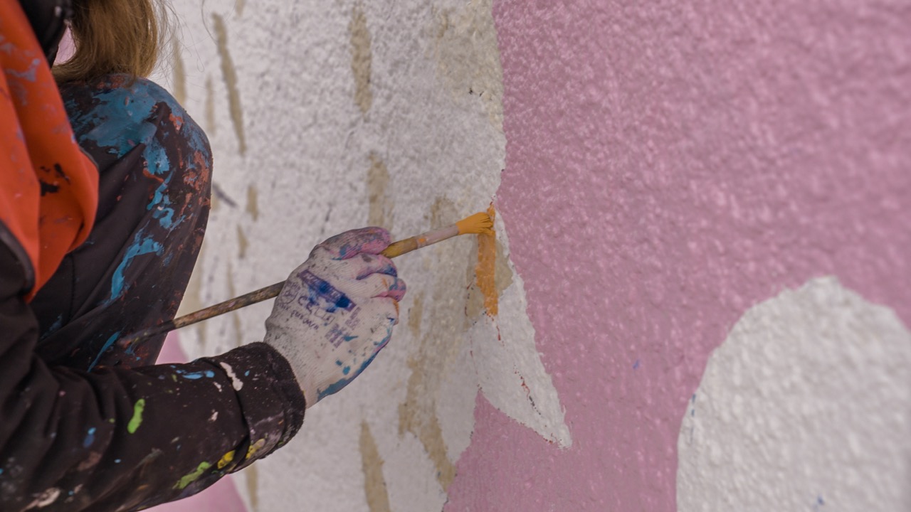 Nahaufnahme einer Hand mit Pinsel beim Ausarbeiten kleiner Details auf einer rosa und weißen Wandgestaltung.
