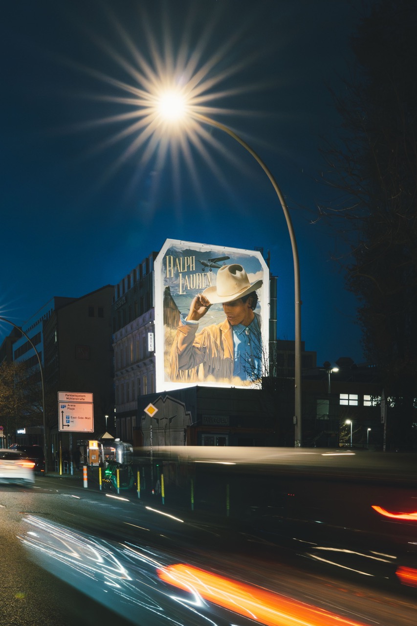 Straßenszene in Berlin mit Passant*innen und Verkehr vor dem großformatigen Ralph-Lauren-Mural.