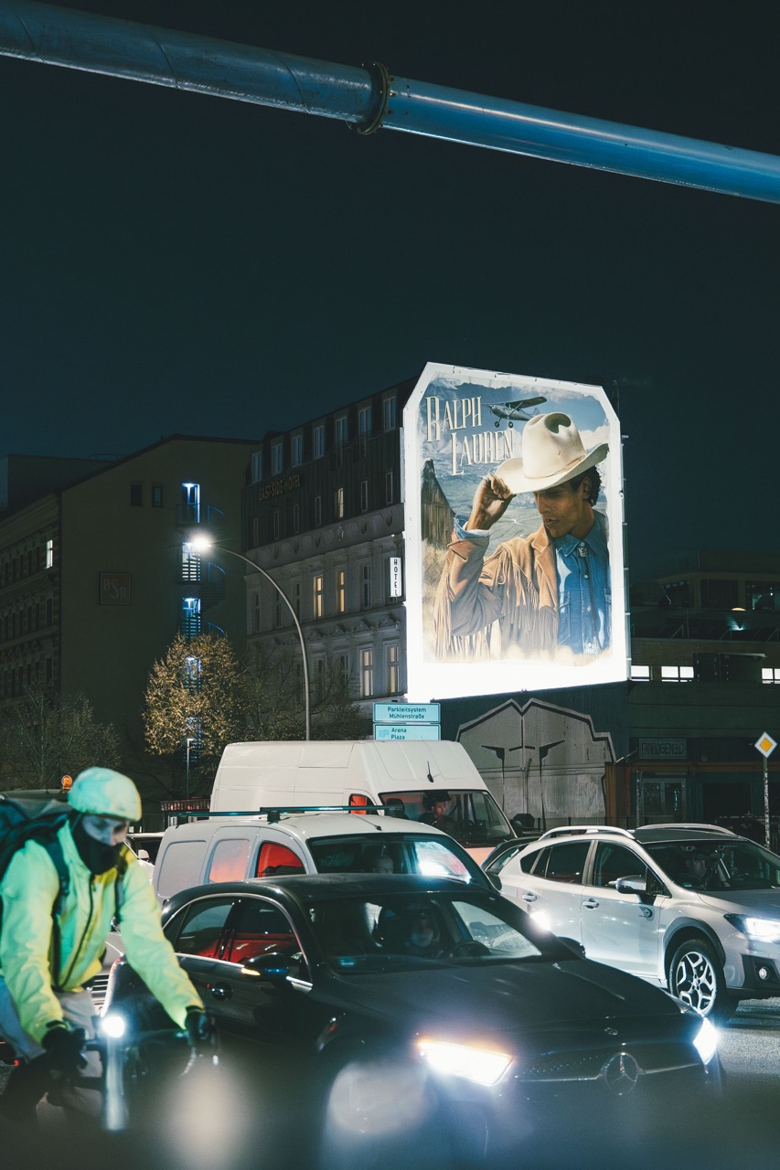 Nächtliche Berliner Straßenszene mit Autos, Fahrradfahrer und großformatigem Ralph-Lauren-Mural im Hintergrund.