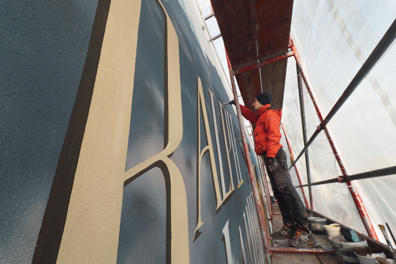 Artist arbeitet auf einem Gerüst an großflächiger Ralph-Lauren-Typografie für ein Mural an der Berliner EAST SIDE GALLERY.