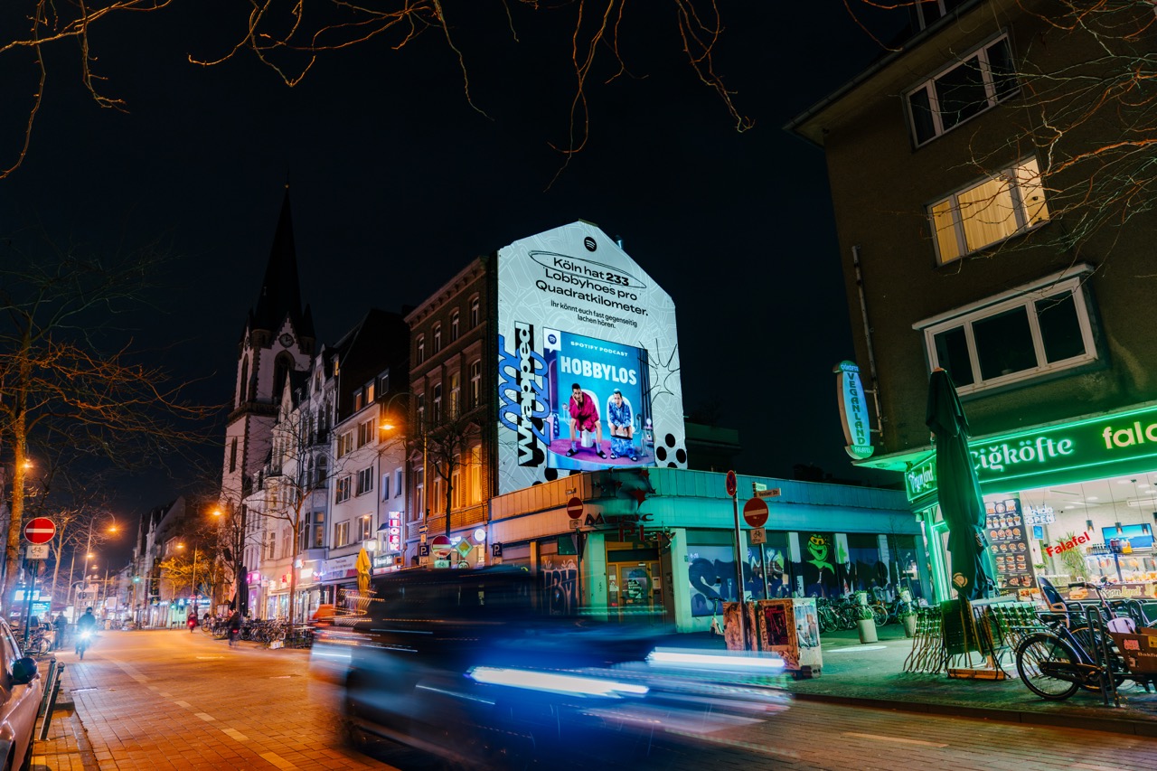 Beleuchtetes Spotify Wrapped Mural mit Podcast-Motiv an einer Kölner Hausfassade bei Nacht.