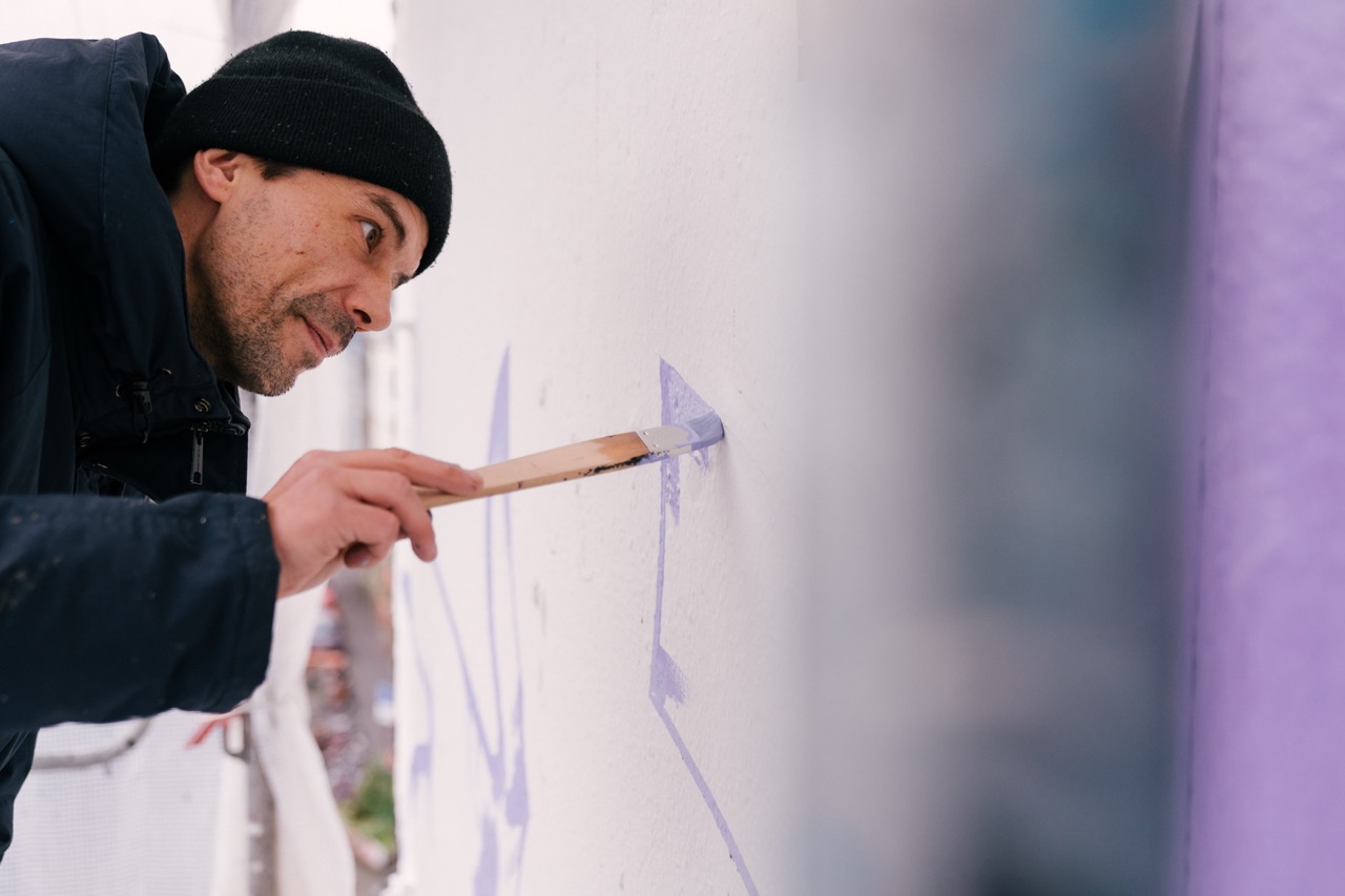 Künstler malt feine lilafarbene Linien und Konturen auf eine Hausfassade während der Entstehung eines Murals.