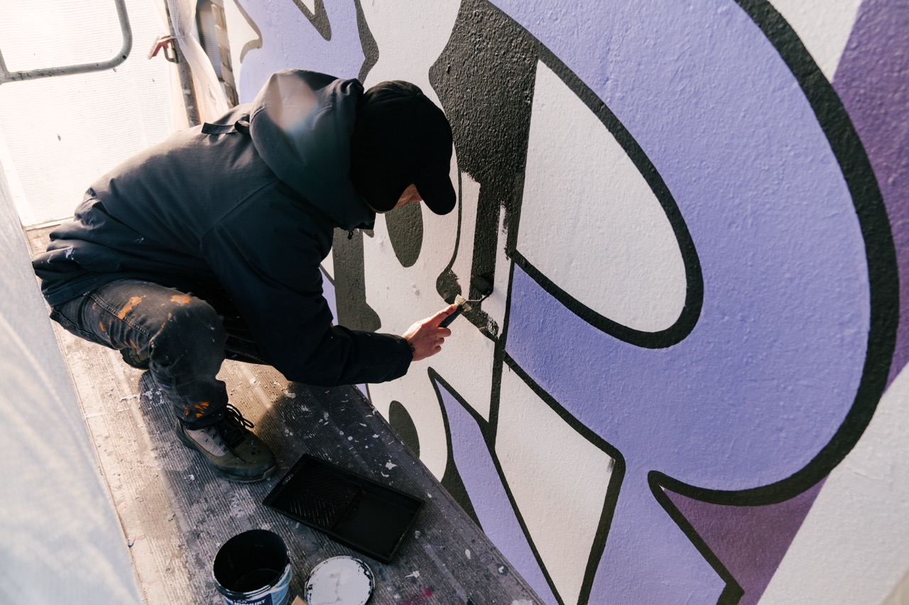 Artist arbeitet mit Farbrolle und Pinsel an großen schwarzen und lilafarbenen Formen eines Spotify Murals.