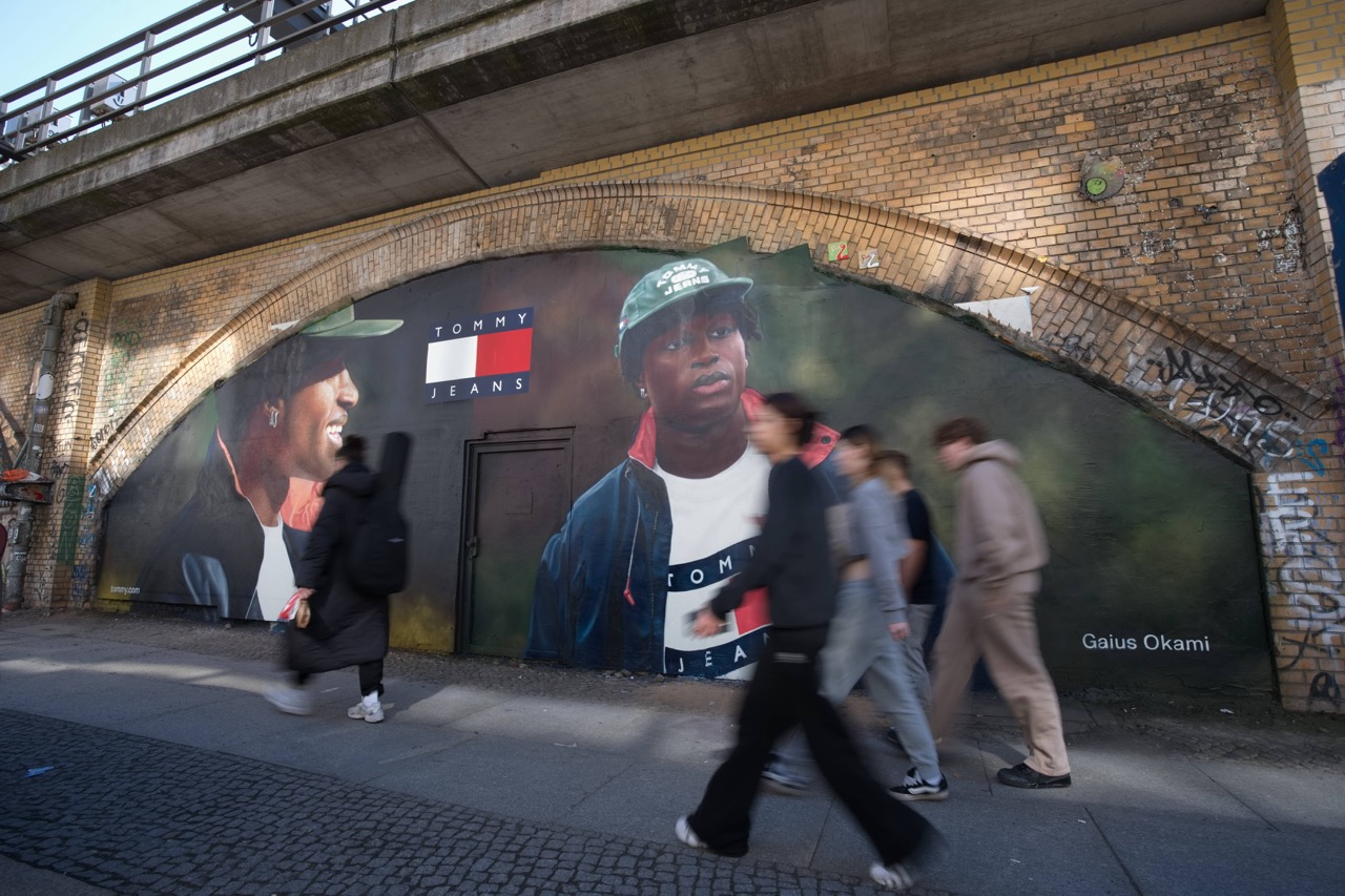 Großformatiges Tommy Hilfiger Mural mit Gaius Okami unter einer Berliner S-Bahn-Trasse, während Passant*innen die Szene durchqueren.