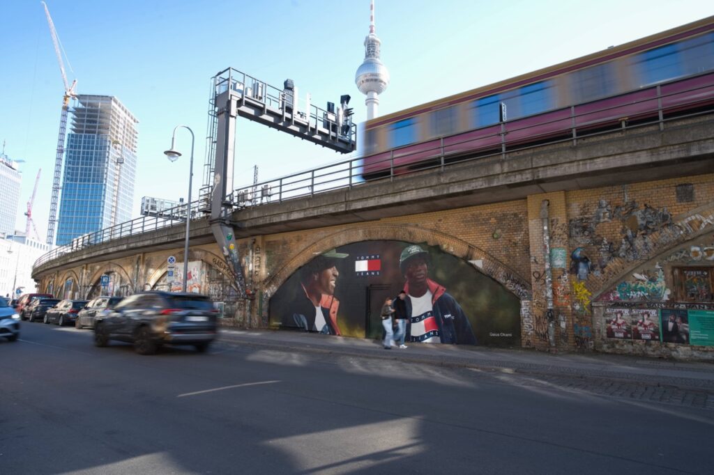 Großformatiges Tommy Hilfiger Mural unter den S-Bahn-Bögen der Diercksenstraße in Berlin-Mitte. Das Artwork zeigt den Künstler und Kreativen Gaius Okami vor urbaner Kulisse, während im Hintergrund eine S-Bahn am Alexanderplatz vorbeifährt.