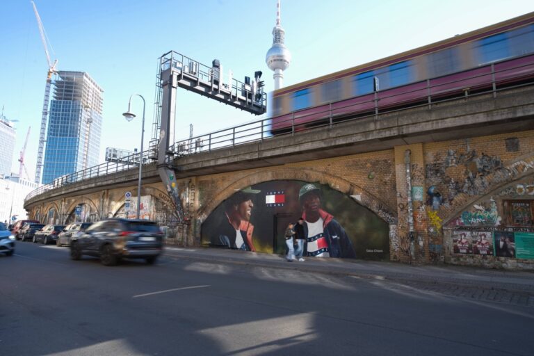 Großformatiges Tommy Hilfiger Mural unter den S-Bahn-Bögen der Diercksenstraße in Berlin-Mitte. Das Artwork zeigt den Künstler und Kreativen Gaius Okami vor urbaner Kulisse, während im Hintergrund eine S-Bahn am Alexanderplatz vorbeifährt.