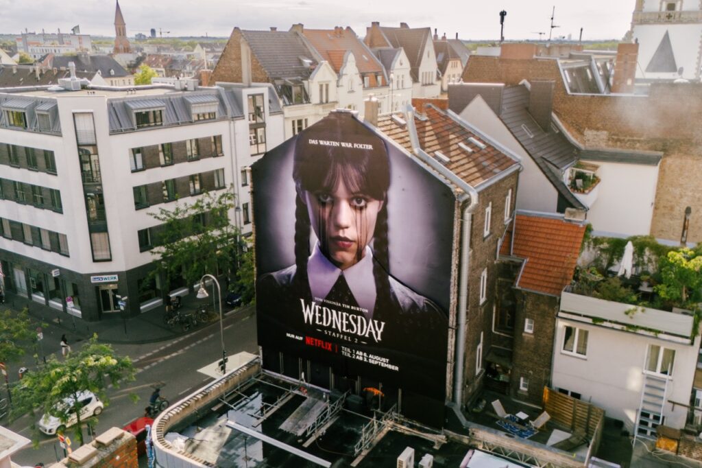 Großformatiges Netflix „Wednesday“-Mural an einer Giebelwand in Köln aus der Vogelperspektive. Die Figur Wednesday blickt frontal in die Kamera, schwarze Tränen verlaufen über ihr Gesicht. Das dunkle Artwork hebt sich stark von der umliegenden Altstadtbebauung ab.