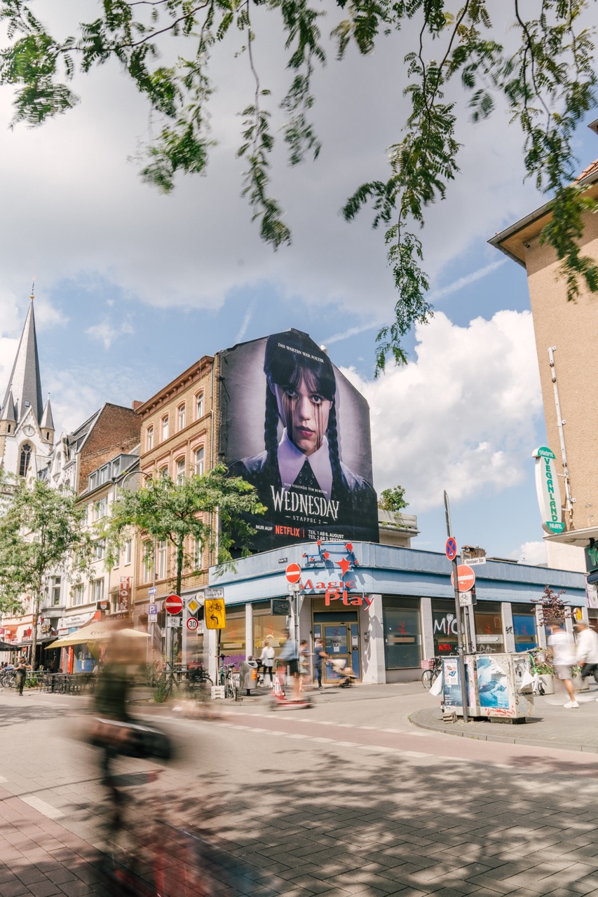 Straßenansicht des fertigen Netflix „Wednesday“-Murals in Köln mit Passant*innen und urbanem Umfeld.